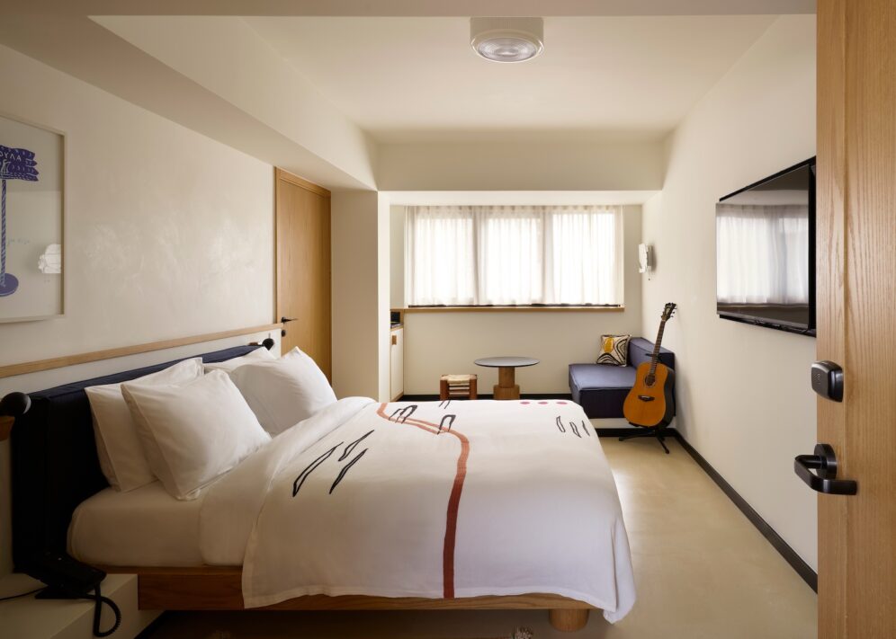 Cozy hotel room with a soft, white duvet and abstract design. A guitar leans against a blue sofa near a window. Warm tones, serene atmosphere.