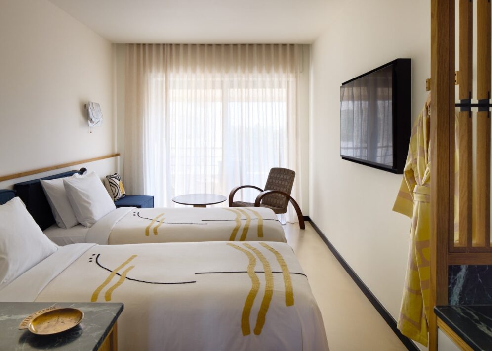 A modern hotel room with two neatly made beds featuring abstract patterned bedspreads. A large window with sheer curtains provides soft natural light. A cozy chair and small round table are near the window, and a wall-mounted TV is opposite the beds, creating a calm, inviting atmosphere.