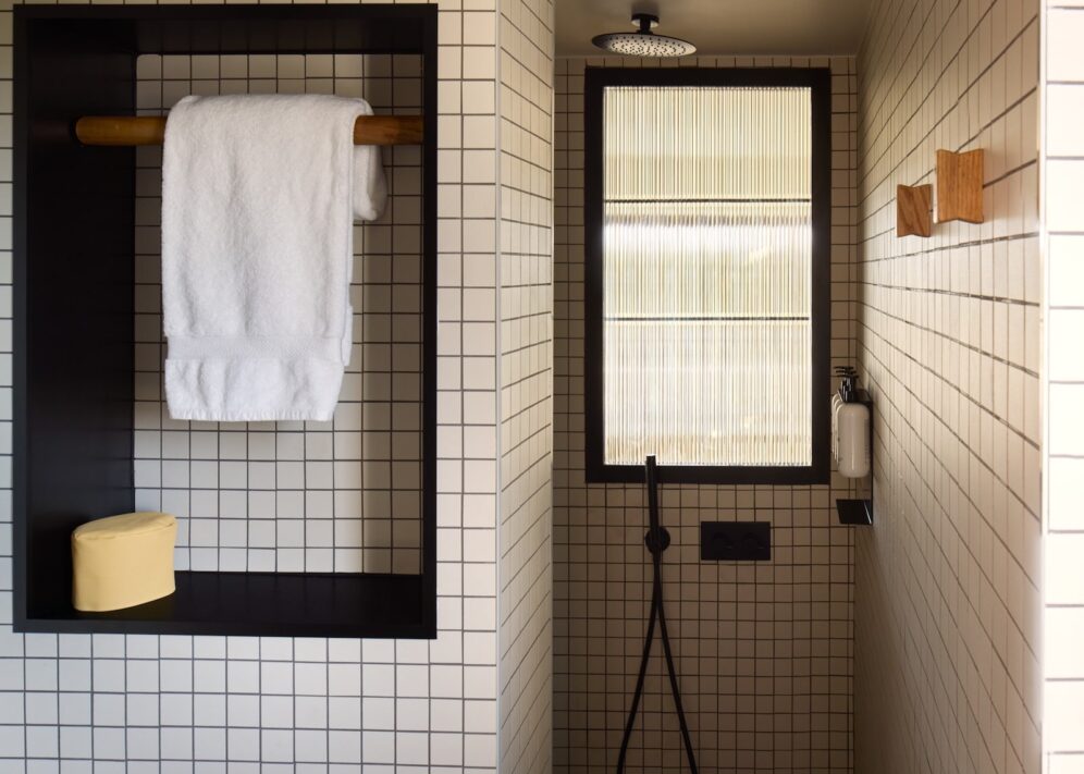 Modern bathroom with grid-patterned tiles, featuring a recessed shelf with a white towel and yellow soap. A rain shower head and a ribbed glass window add elegance.