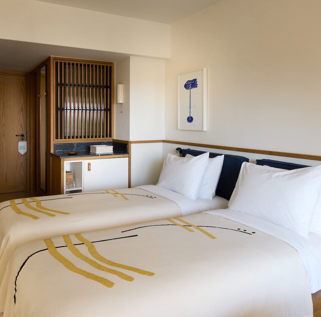 Modern hotel room with two neatly made single beds adorned with abstract-patterned blankets. Warm wooden accents, a small art piece, and soft lighting create a cozy atmosphere.