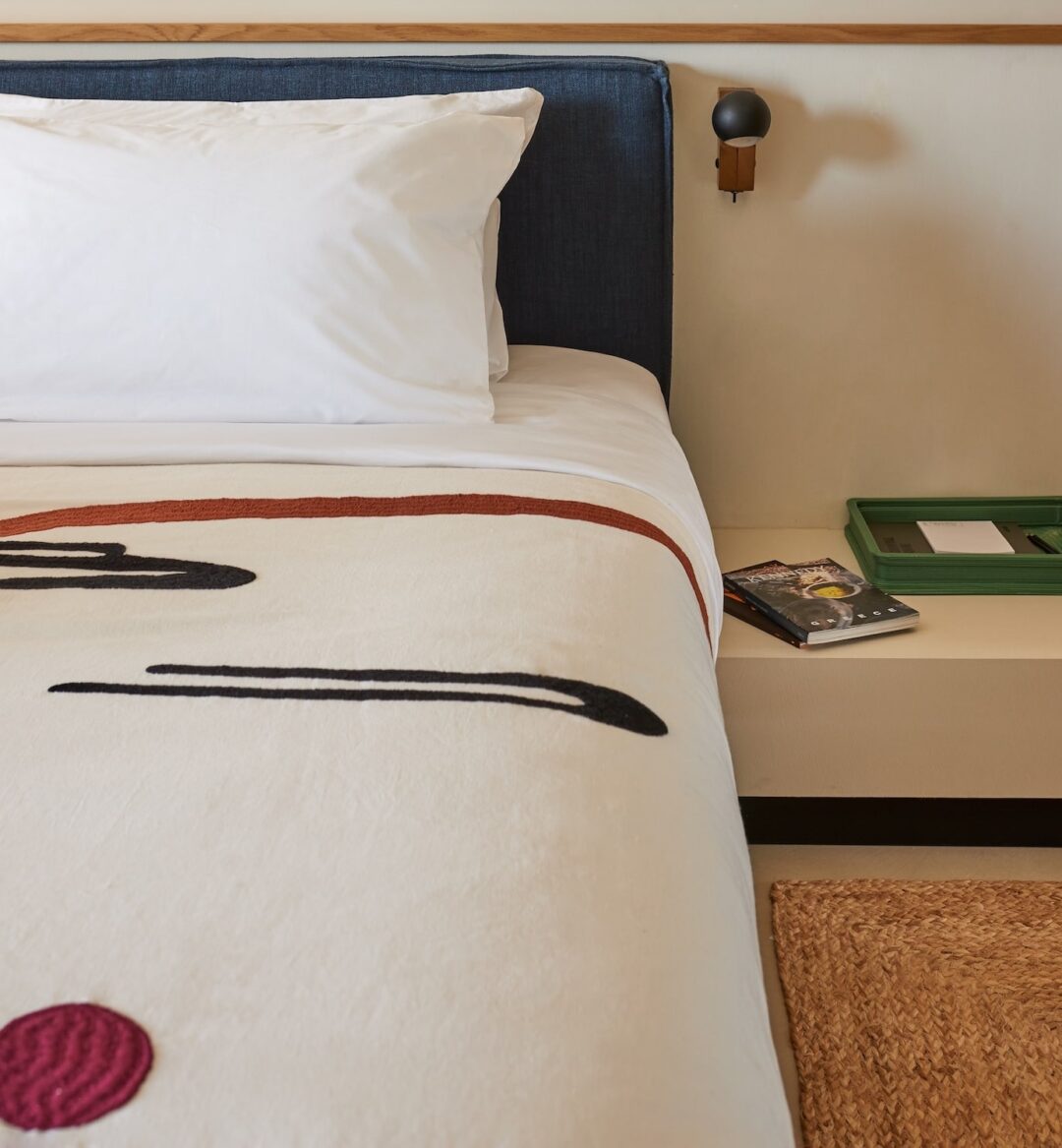 A neatly made bed with a white pillow and a blanket featuring abstract black and red lines. A nightstand holds a green tray and a book, creating a minimalist and calm atmosphere.