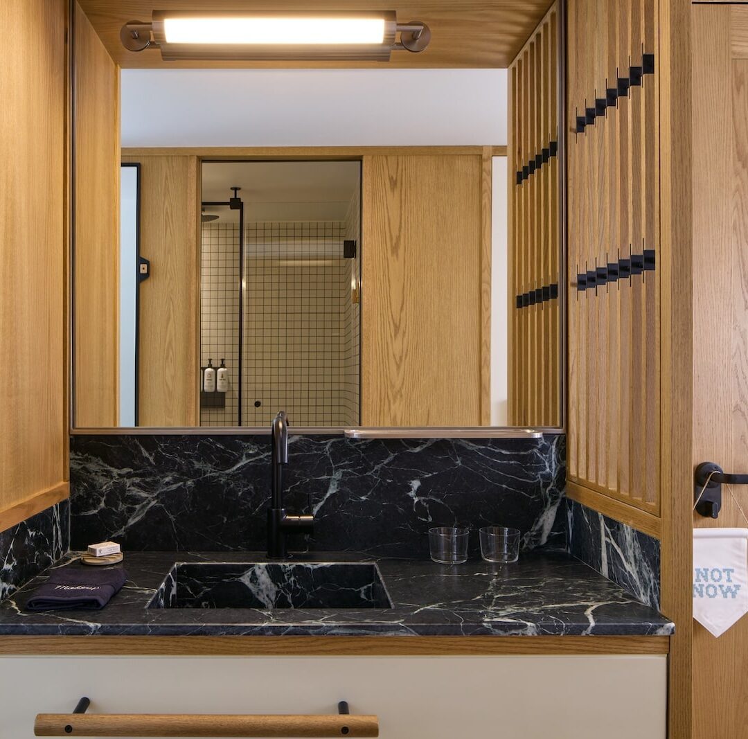 Modern bathroom with a black marble sink, wooden walls, and a rectangular mirror reflecting a shower with tiled walls. Warm, minimalist design.