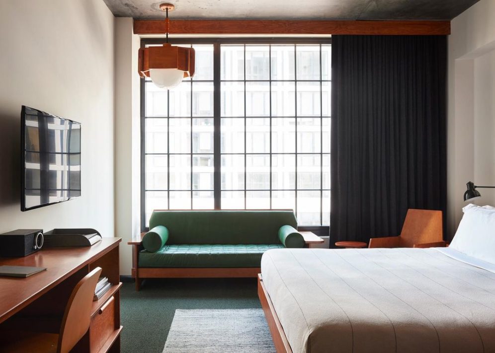 Room at Ace Hotel featuring a bed with white linen, a big ceiling height window, green leather sofa and a table with a TV