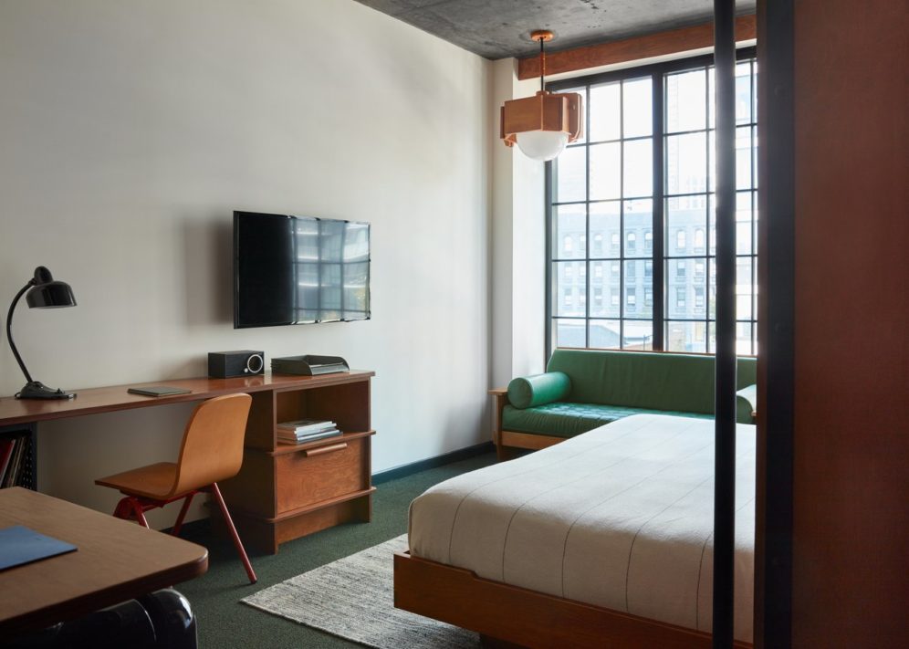 Queen room in Ace hotel featuring a bed, green leather sofa, a table with a chair, a TV, a big window with a city view