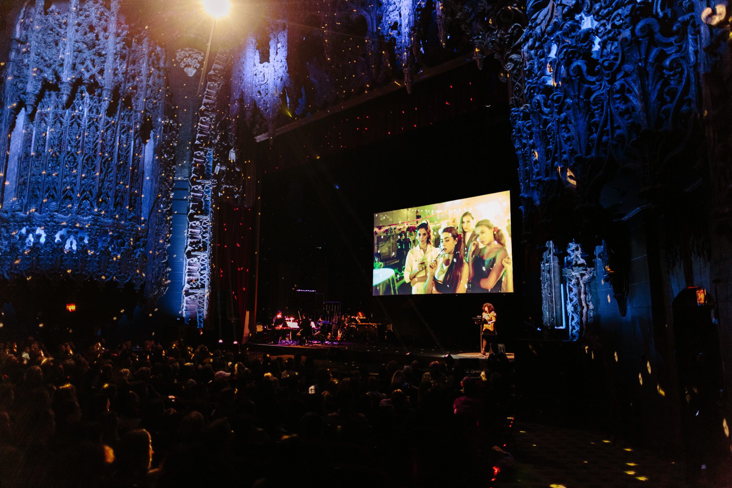 Image of Pop-Up Magazine at The Theatre at Ace Hotel in Los Angeles, CA. In the center of the image you see an image projected on the screen, a person standing on a podium on the right side of the stage, with blue and purple lights on the proscenium.