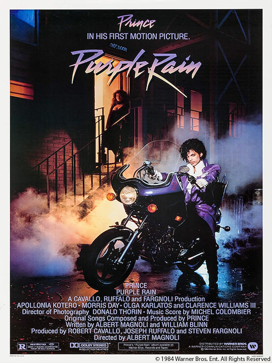 Text: Prince IN HIS FIRST MOTION PICTURE "PURPLE RAIN" Pictured: Prince on a motorcycle in an alleyway. Behind him is Apollonia Kotero in a door way.