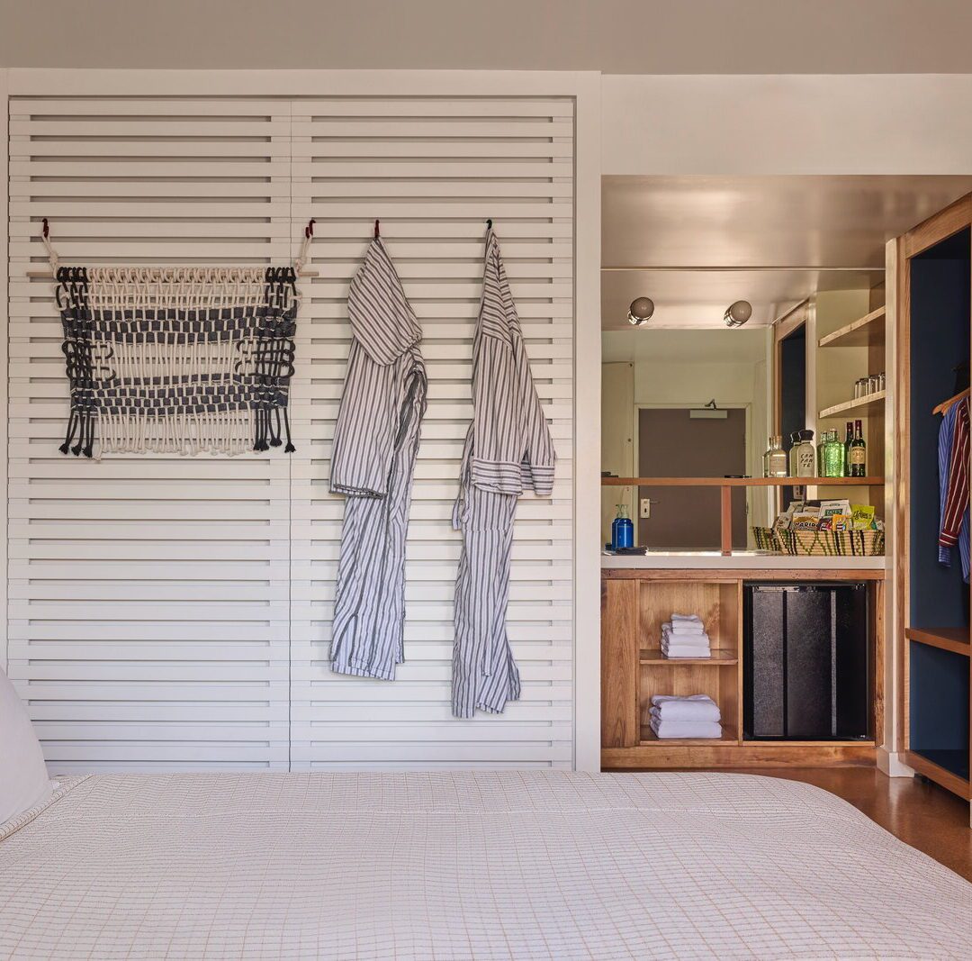 two robes hang against a slotted wall, and a large mirror sits above towels and a mini fridge