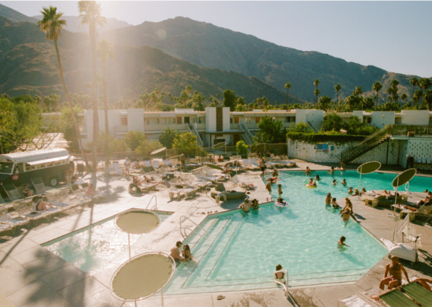 Scenic view of the pool area at Ace Hotel & Swim Club, set against a backdrop of towering palm trees and rugged mountains. Guests are lounging on sunbeds and swimming in the clear, inviting water. The sun is shining brightly, casting a warm glow over the entire scene, creating a perfect summer day atmosphere.