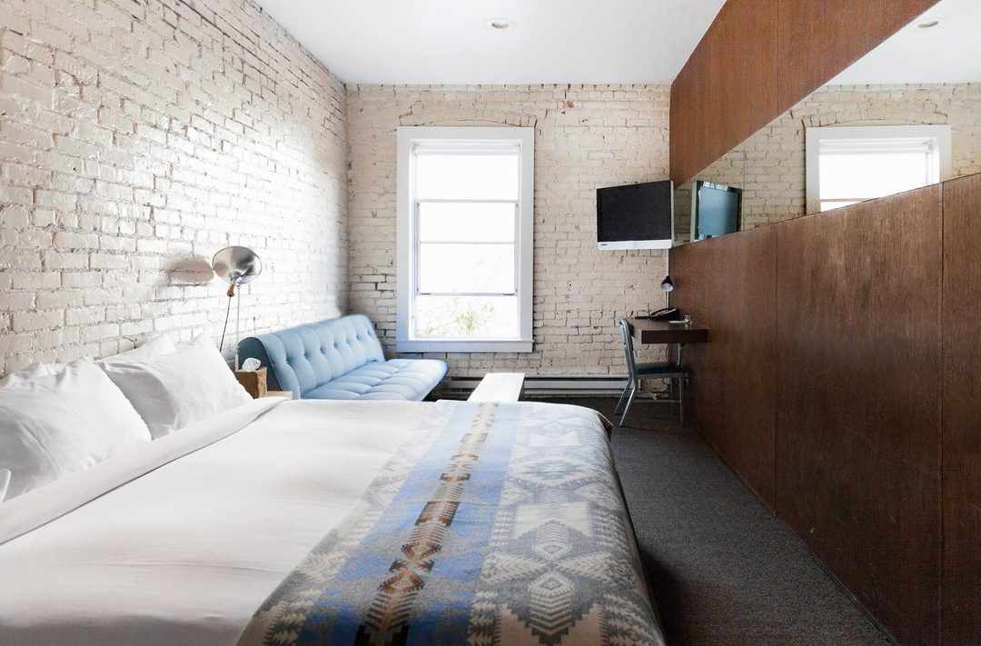 a room with exposed white brick walls is drenched in natural sunlight; a bed sits next to a long couch