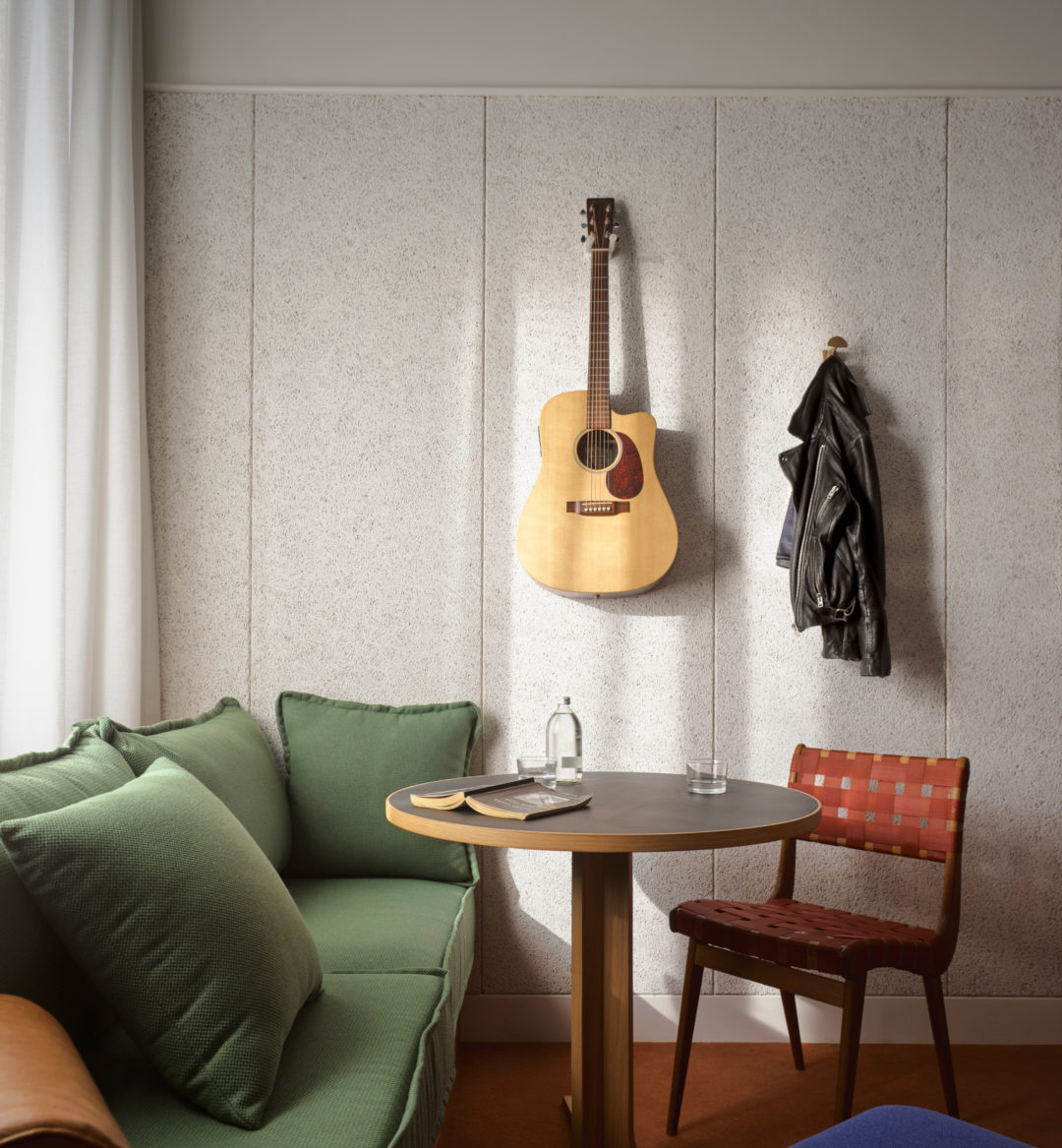 Green sofa and throw pillows on one side, red chair on the other surrounding small round table, with guitar hanging on wall in background