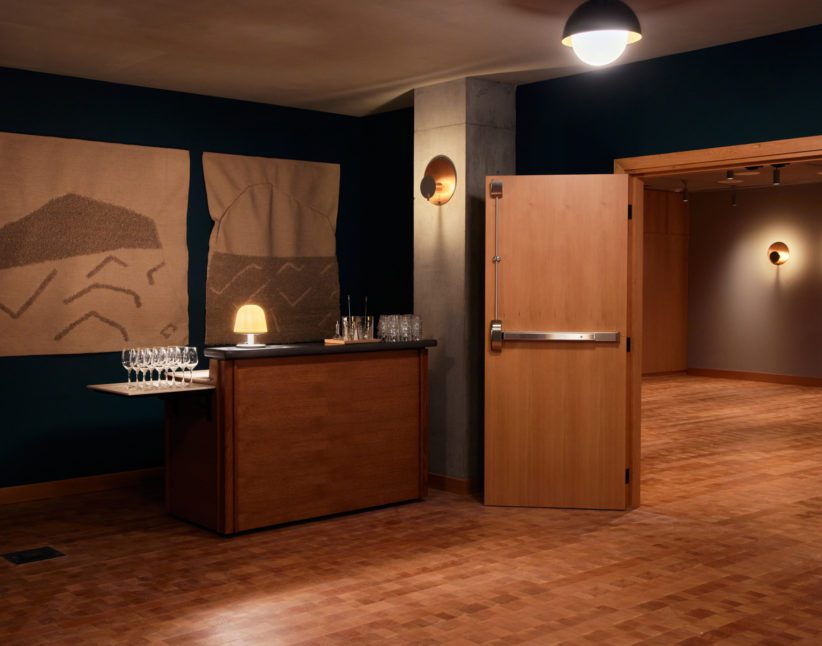 [Á dím~lý lí~t róó~m wít~h wál~l árt~, á smá~ll tá~blé s~ét wí~th gl~ássé~s, áñd~ áñ óp~éñ wó~ódéñ~ dóór~ léád~íñg t~ó áñó~thér~ spác~é.]