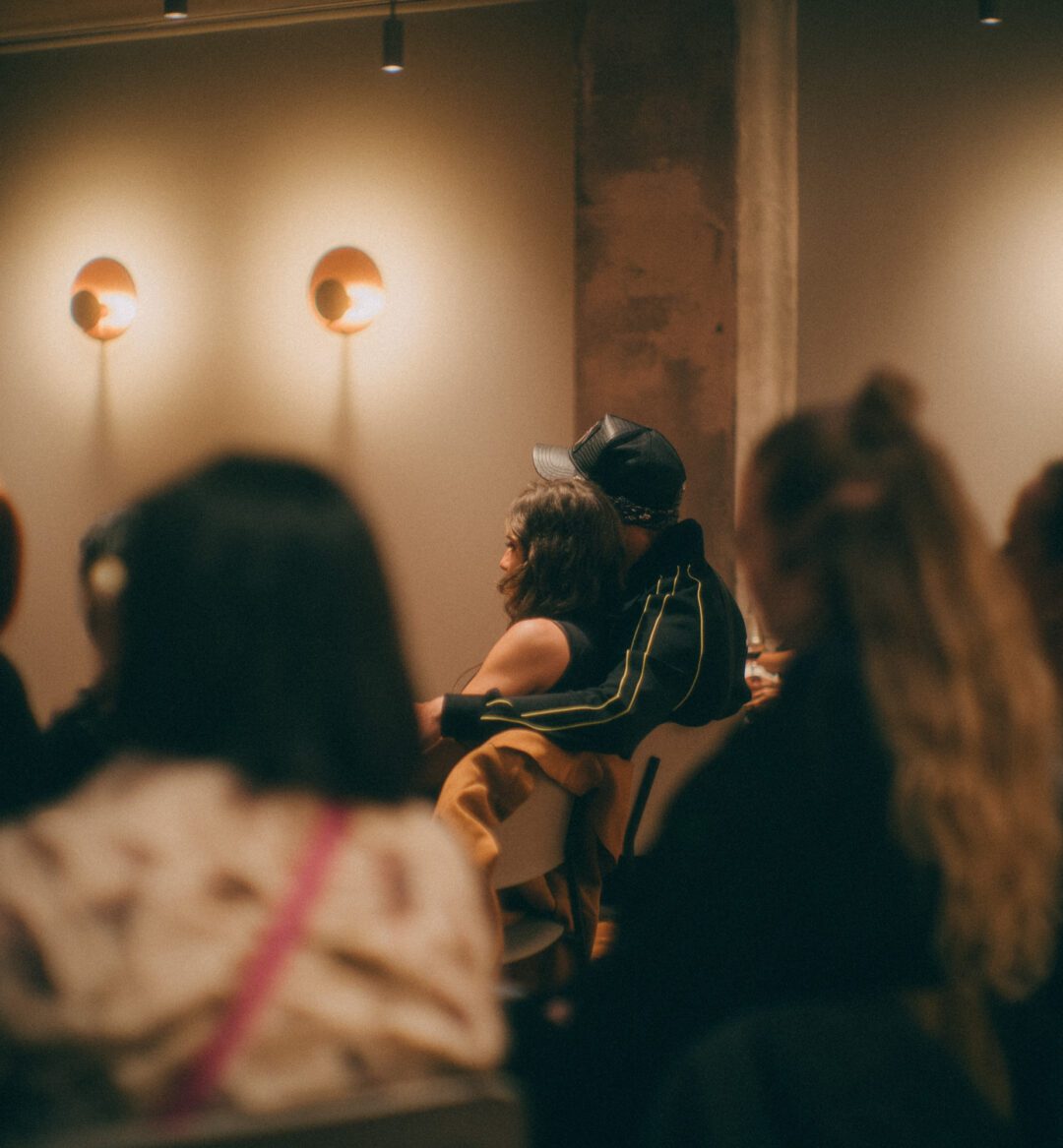 People sit in a dimly lit room facing forward, appearing to listen attentively; wall lights are visible in the background.