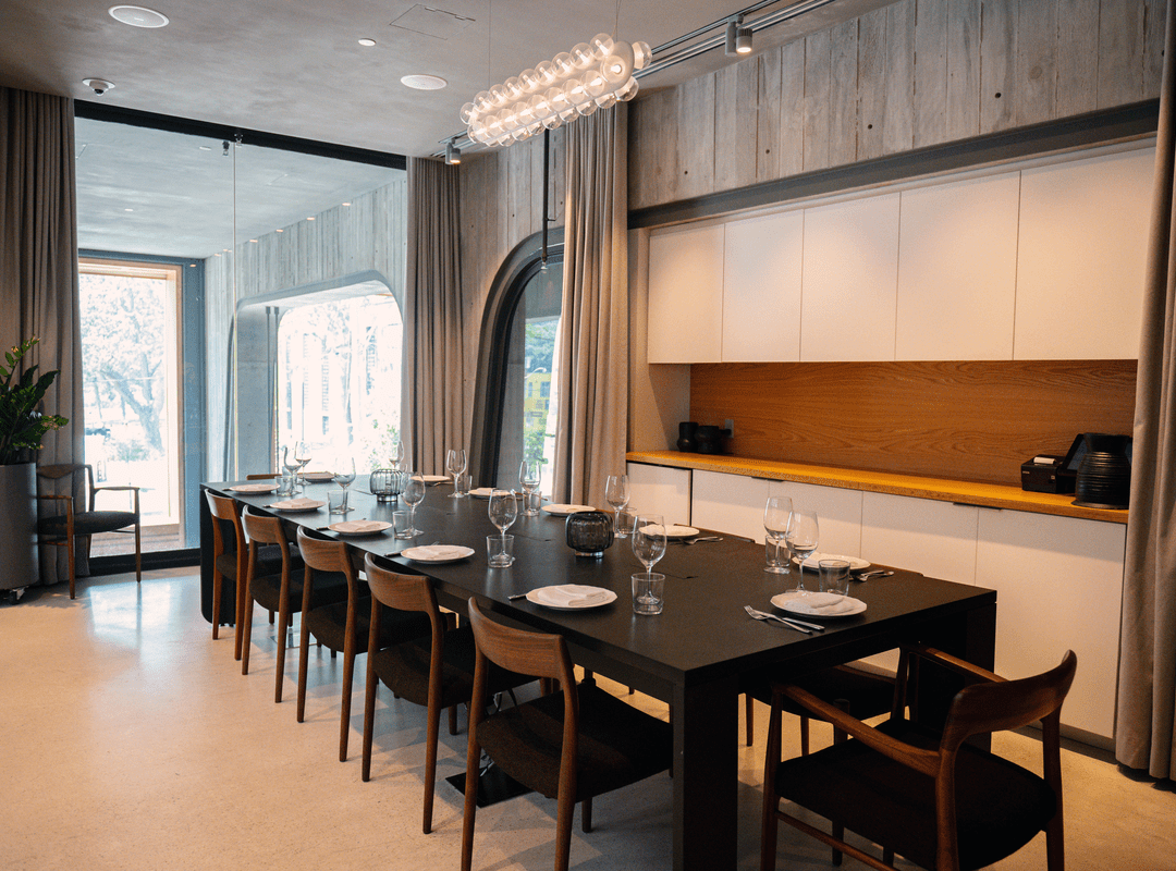Modern dining room with a long black table set for eight, wooden chairs, large windows, and minimalist decor.