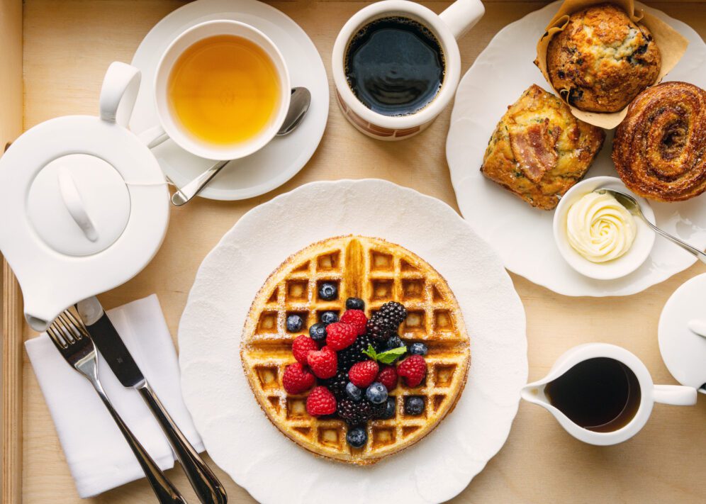 A delicious breakfast spread featuring a golden waffle topped with fresh berries, a cup of black coffee, a pot of tea, a selection of pastries including a muffin and a croissant, and a small dish of butter, all beautifully arranged on a white plate with cutlery on a napkin. Ace Hotel Toronto