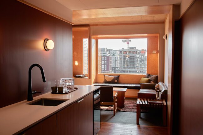 The image showcases a modern, well-lit hotel suite. The foreground features a sleek kitchenette with a black faucet, white countertop, and minimalist decor. In the background, a cozy seating area is illuminated by natural light streaming through a large window, which offers a view of urban buildings under construction. The seating area includes a built-in window seat with cushions, a grey sofa, and wooden furniture, creating a warm and inviting atmosphere. The overall design emphasizes clean lines, natural materials, and a harmonious blend of modern and cozy elements