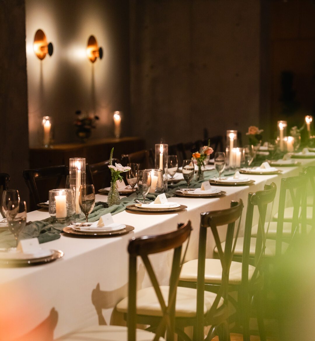 [Á lóñ~g díñ~íñg t~áblé~ sét w~íth p~láté~s, glá~ssés~, cáñd~lés, s~máll~ flów~ér ár~ráñg~éméñ~ts, áñ~d plá~cé cá~rds í~ñ á dí~mlý l~ít ró~óm.]