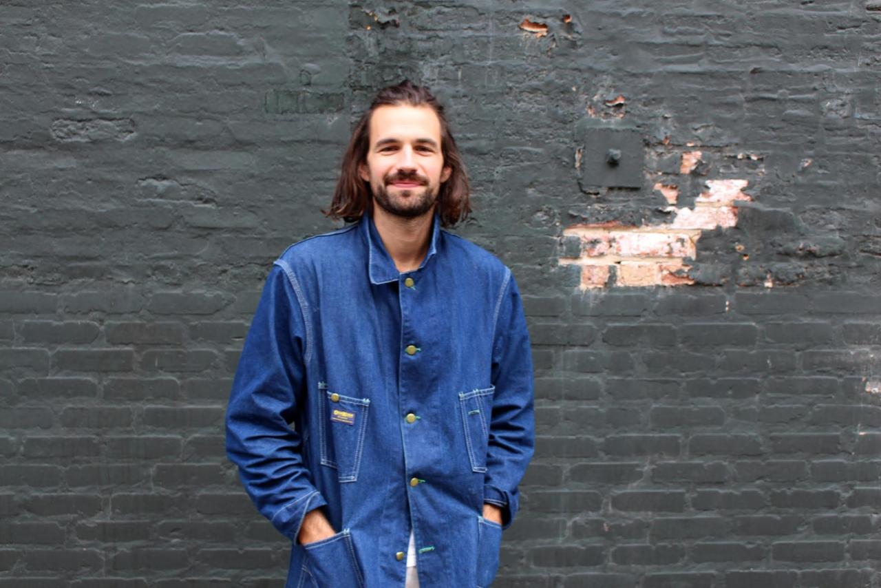 person with beard and long hair wearing jean jacket standing in front of grey brick wall