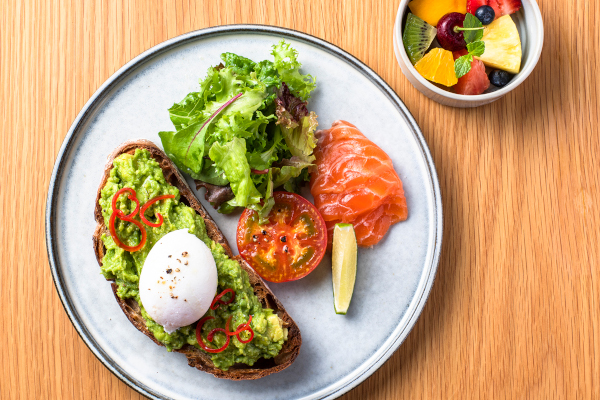 White plate with avocado toast and poached egg, with tomato salad on side, on bright beige tabletop. Small bowl of fruit in the upper right corner